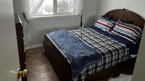 A bedroom with a bed, a window with sheer curtains, a wooden headboard, and a door with a brass doorknob. - Cozy Country Living (Nipawin)