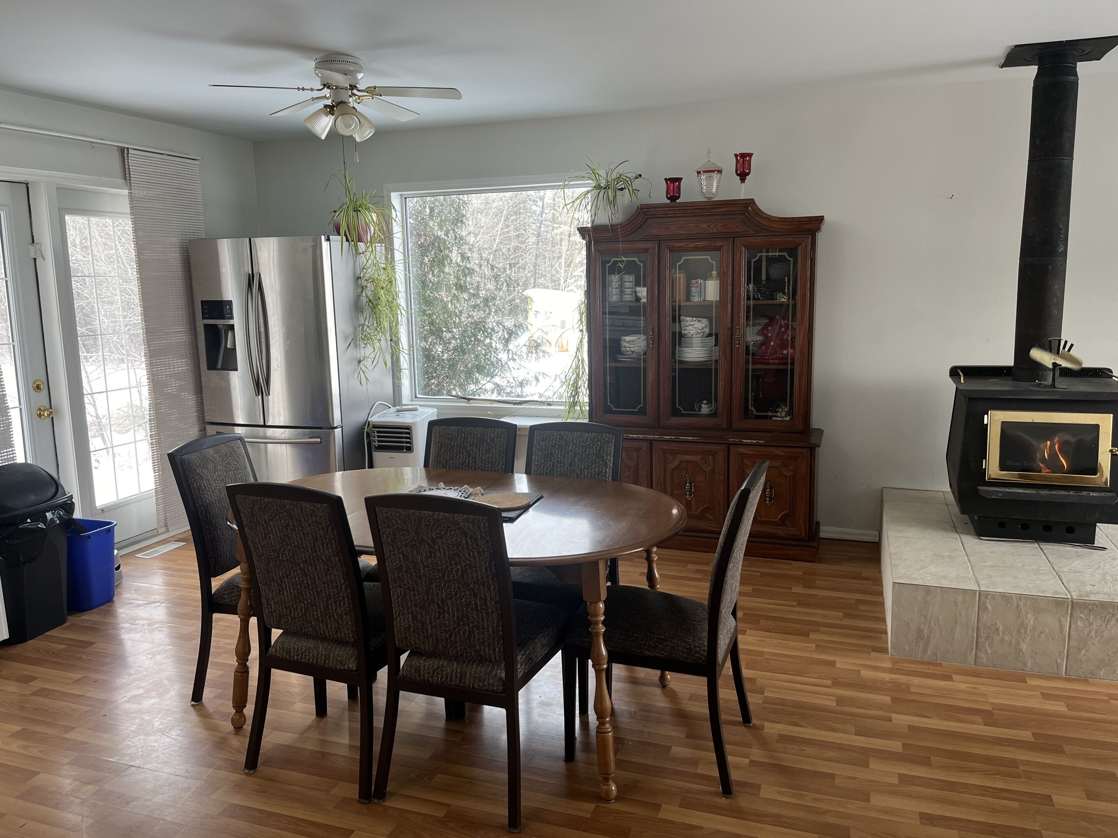 A dining area with a round table and six chairs, a wooden cabinet with glass doors, a black wood-burning stove, and a ceiling fan.