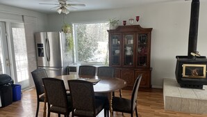 A dining area with a round table and six chairs, a wooden cabinet with glass doors, a black wood-burning stove, and a ceiling fan. - Cozy Country Living (Nipawin)