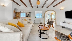 A spacious living area with a sofa, coffee table, and chairs, a kitchen in the background, and a TV on the right. - Villa Adriana with Sea View, Mountain View, Wi-Fi and Air Conditioning (Dénia)