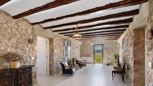 Interior com paredes de pedra, teto de madeira, lareira e área de estar com duas cadeiras e uma mesa.