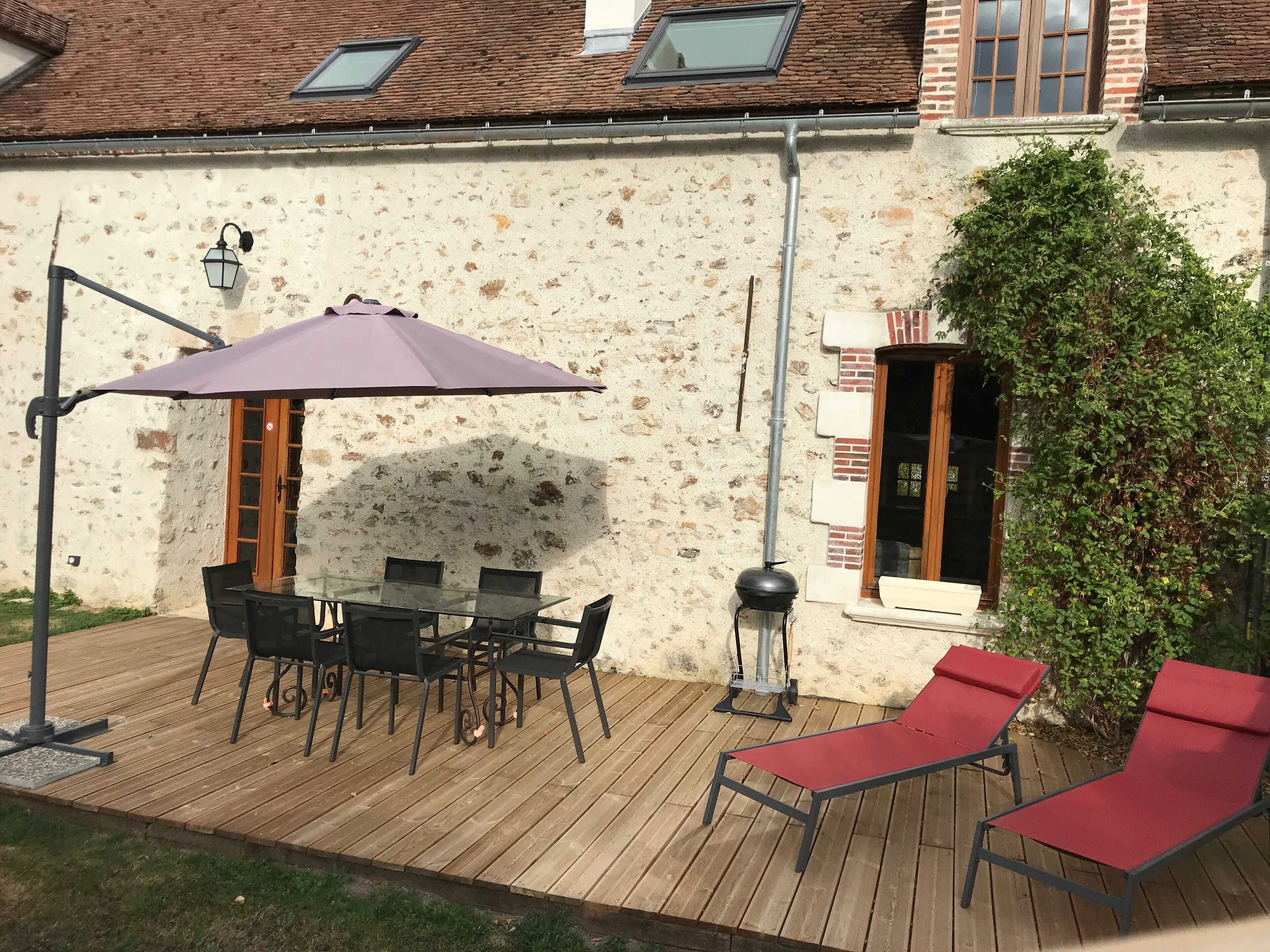 An outdoor patio with a table and chairs, a red lounge chair, a purple umbrella, and a brick wall with a window.