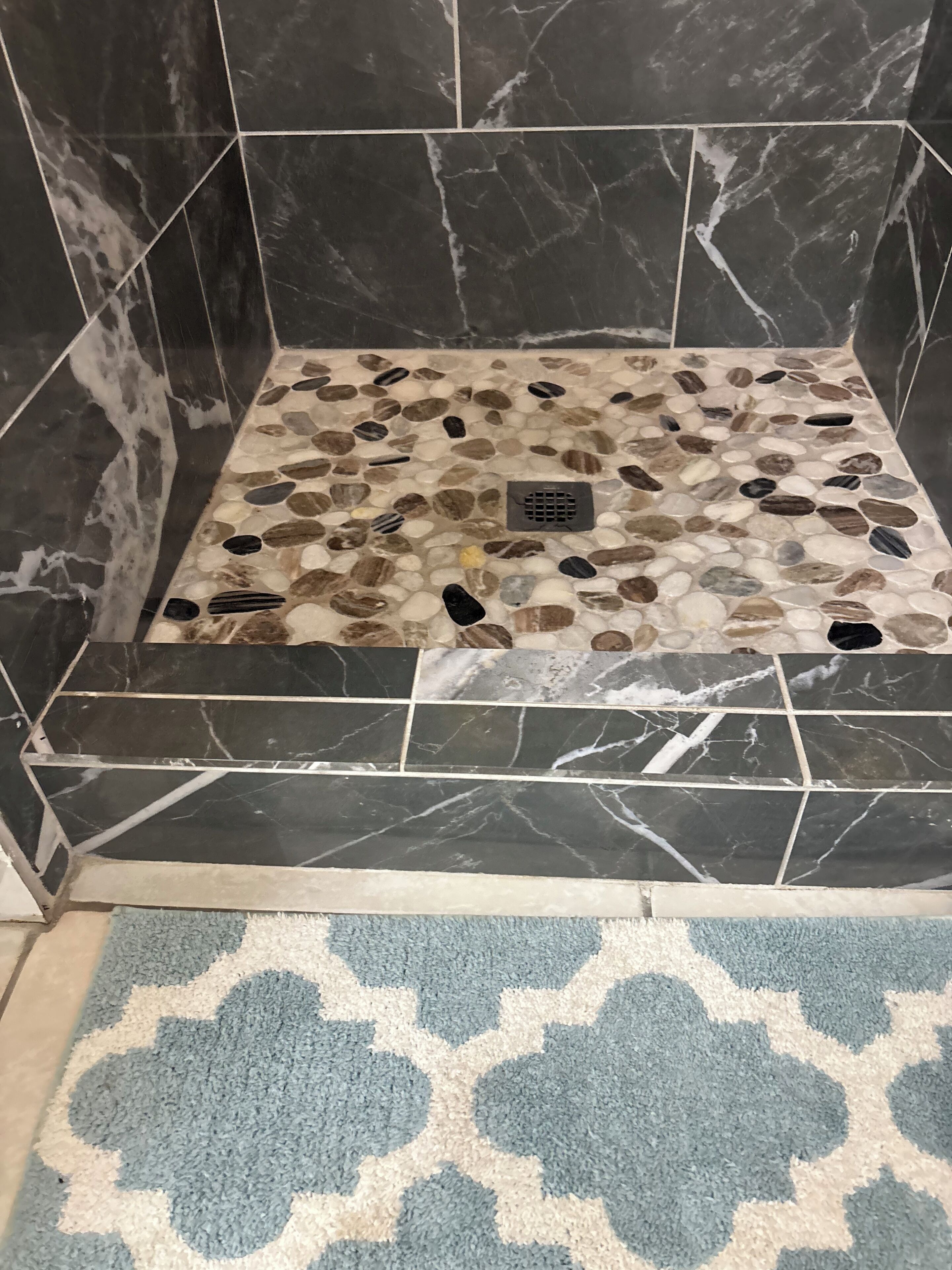 A shower area with a pebble mosaic floor, black marble tiles, and a patterned rug.
