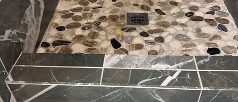 A shower area with a pebble mosaic floor, black marble tiles, and a patterned rug.