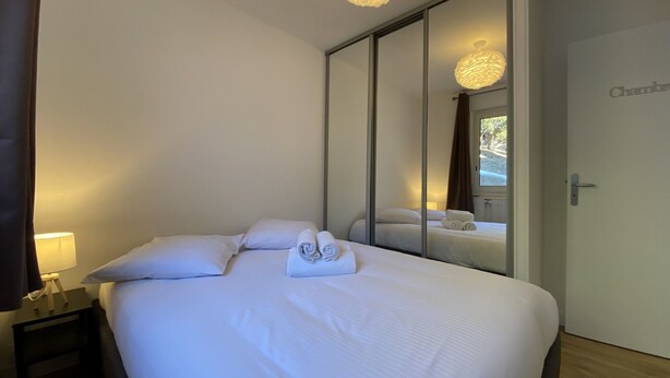A hotel room with a large bed, a bedside table with a lamp, a mirror, and a door labeled "Chambre".