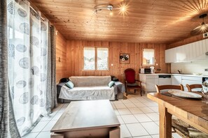 Interior de una cabaña de madera con sofá, mesa de comedor, zona de cocina y vistas al exterior.