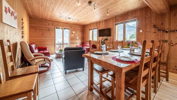 A wooden interior with a dining table set for four, a sofa, and a television mounted on the wall.