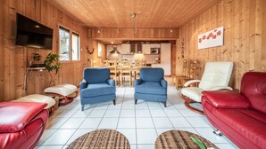 A wooden interior with a TV, two blue armchairs, a red sofa, and a kitchen area.