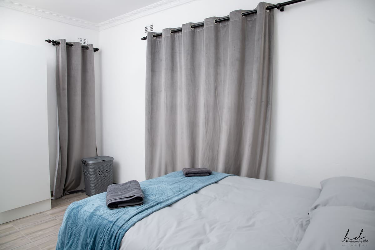 A bedroom with a bed, grey curtains, a grey trash bin, and a blue blanket.