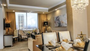 A hotel room with a dining area, a sofa, a TV, and a large window with a city view.
