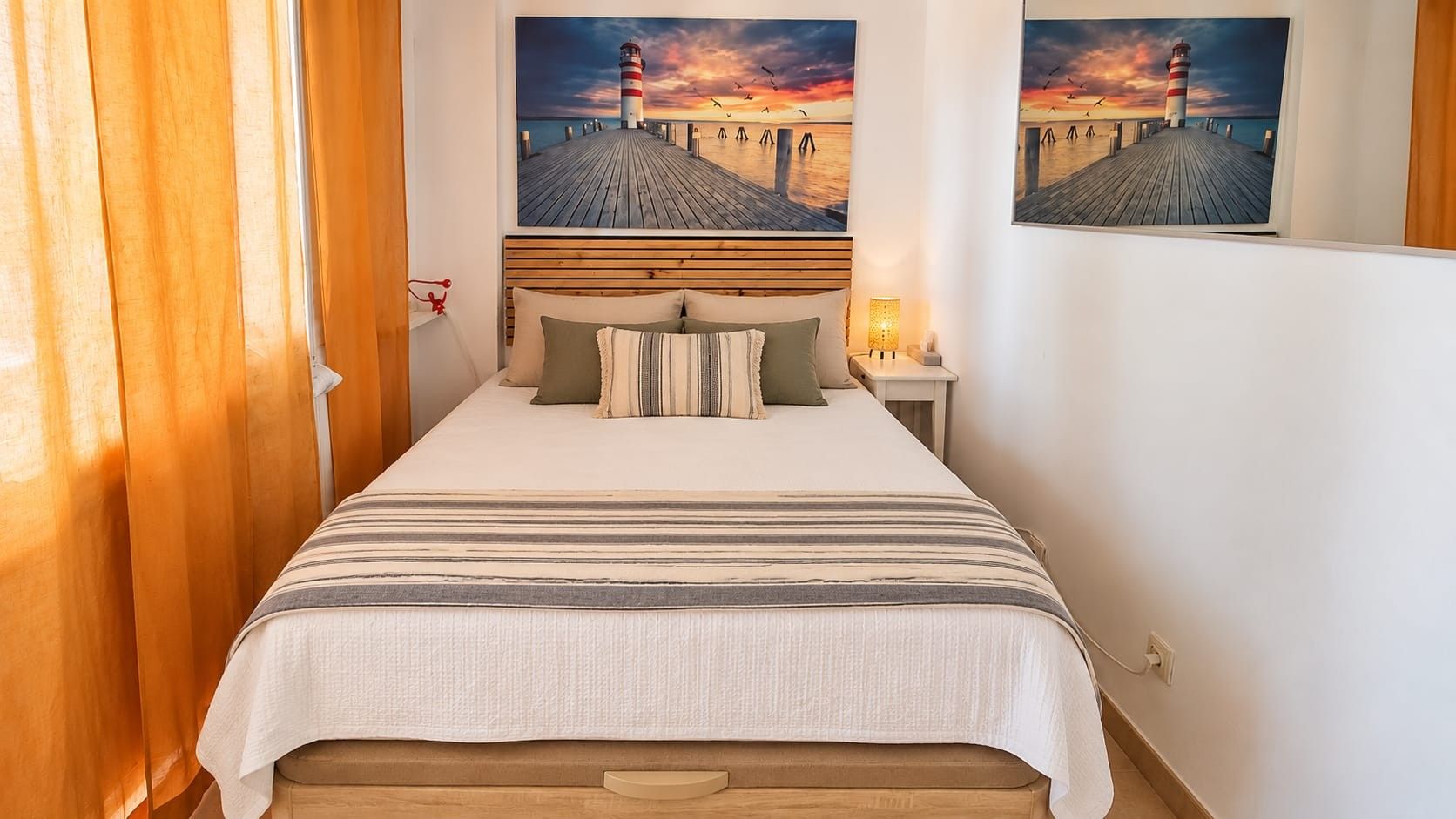 Un dormitorio con cama, almohadas, una mesita de noche con lámpara y dos cuadros colgados en la pared de un faro.