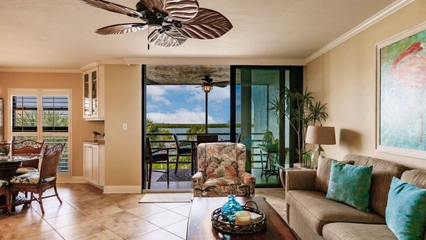 A spacious living room with a large window, a ceiling fan, a dining area with chairs, and a sofa with cushions.