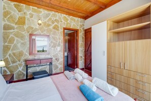 A bedroom with stone walls, a wooden ceiling, a bed, a desk, a wardrobe, and a bathroom door.