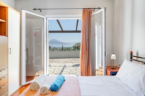 A bedroom with a bed, a nightstand, a lamp, a window with curtains, and a view of the outdoors.