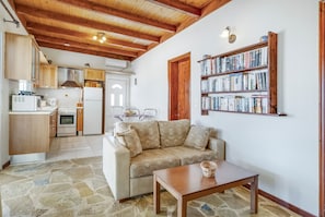 A cozy living area with a sofa, a wooden table, a bookshelf, and a kitchen in the background.