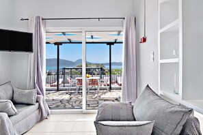 A modern living room with a sofa, a flat-screen TV, and a sliding glass door leading to an outdoor patio with a view of the sea and mountains. - Villa Jimmy's Katomeri 2 with Sea View, Wi-Fi and Air Conditioning (?eganisi)