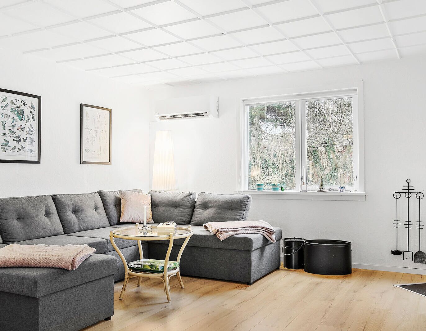 Un salon moderne avec un canapé d’angle gris, une table basse en verre et une petite chaise en bois. Une fenêtre offre une vue sur les arbres, et un présentoir en métal noir se trouve sur la droite.