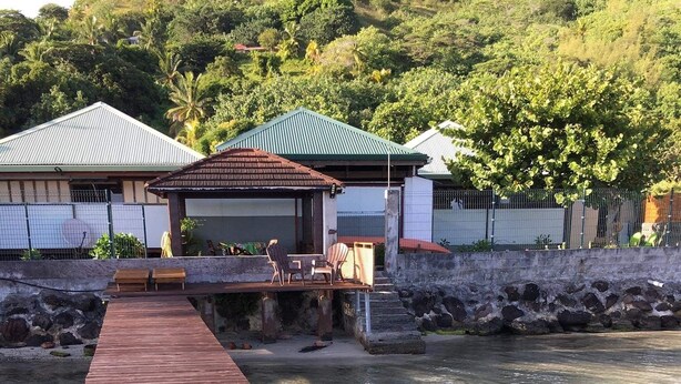 A wooden pier leading to a small outdoor seating area with chairs and a table, overlooking a body of water with clear water and a green hillside in the background.