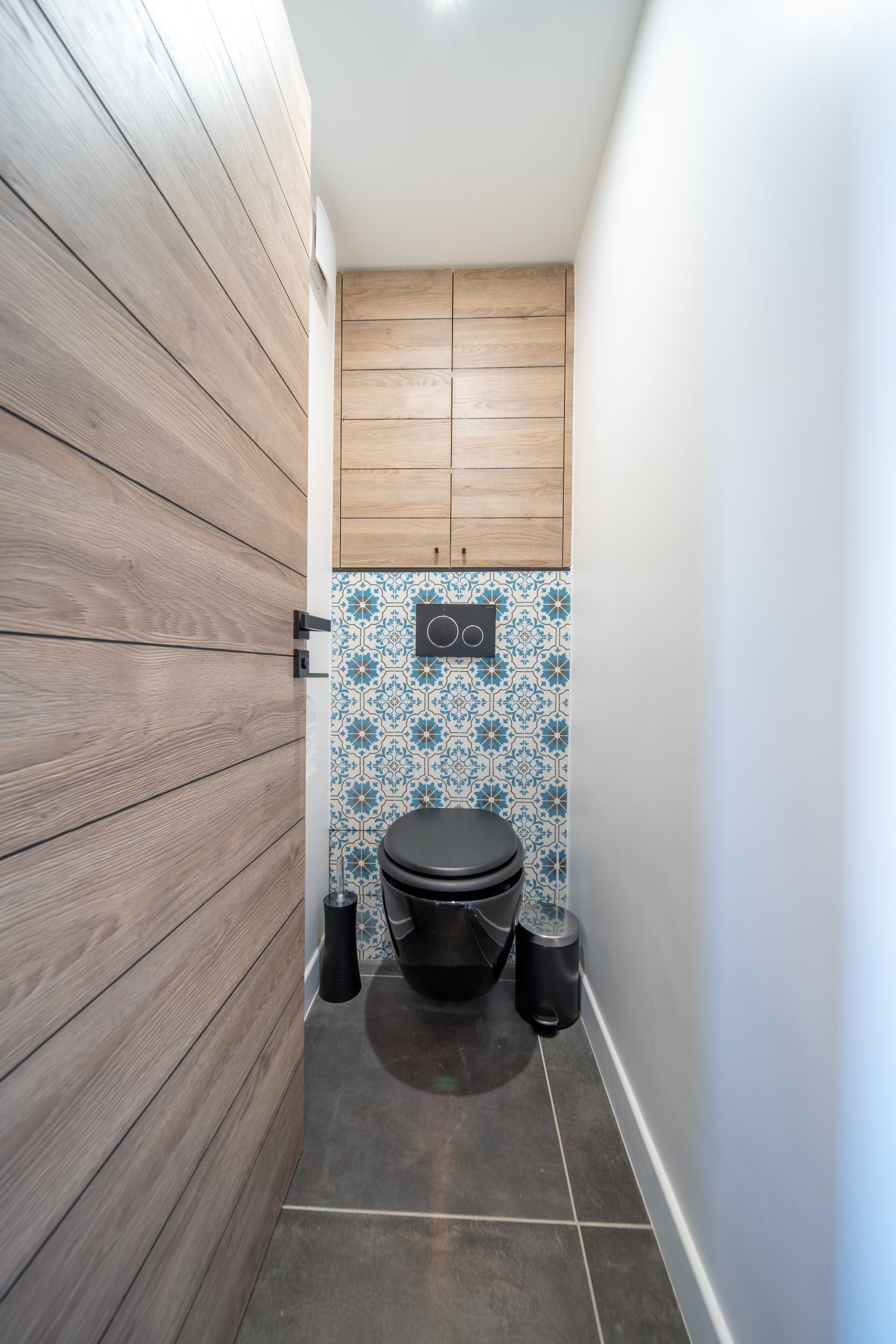 Une petite salle de bain avec un mur lambrissé en bois, des toilettes et un lavabo doté d’un pare-éclaboussures à motifs bleus.