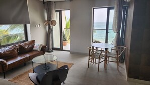 Un salon moderne avec un canapé en cuir, une table basse en verre et un coin repas avec des chaises en bois. Une grande fenêtre offre une vue sur l’océan.