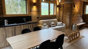A modern cabin interior with a wooden dining table, black chairs, a sofa, and a kitchen area with a large window.