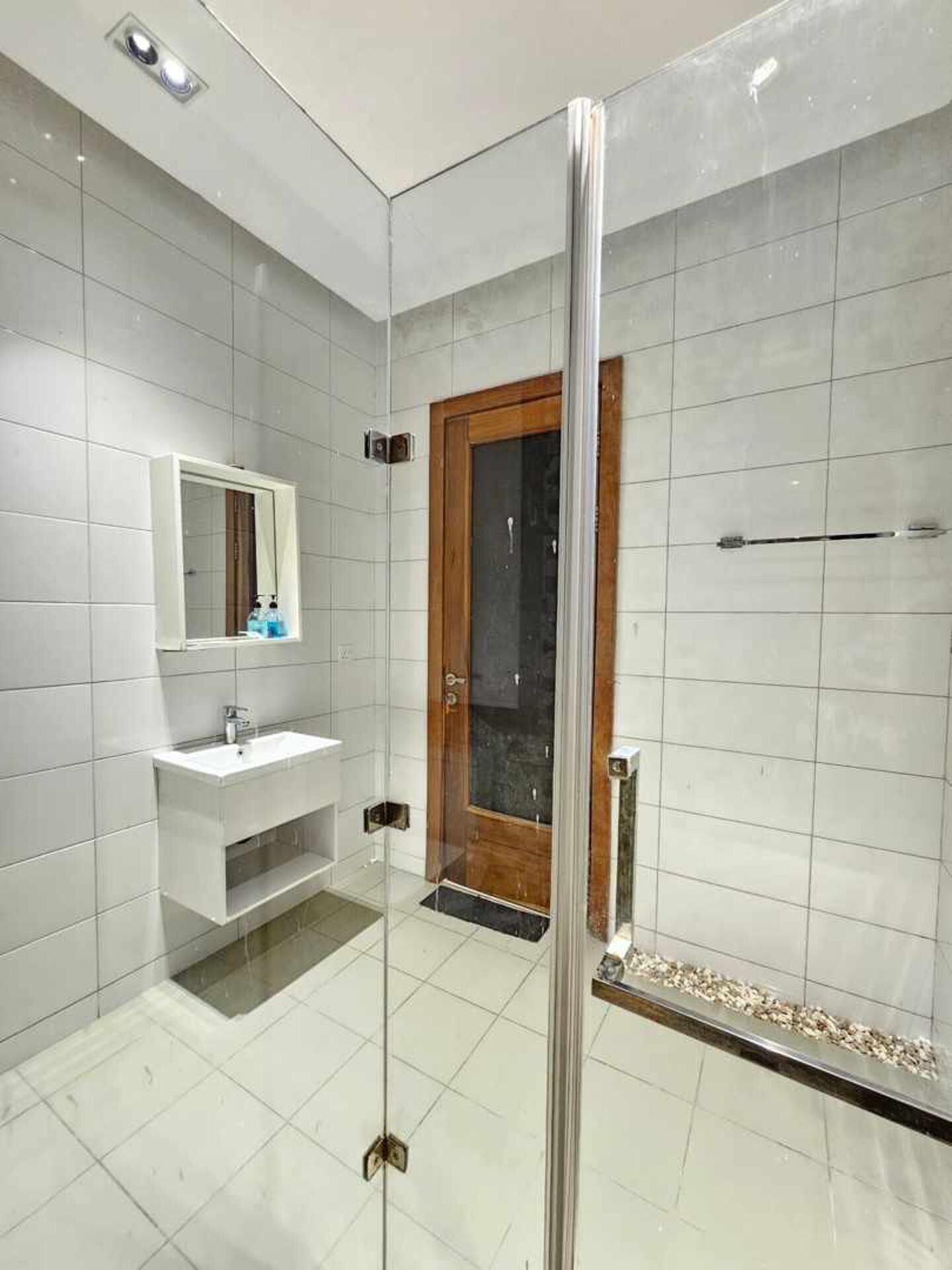 A modern bathroom with a glass-enclosed shower, a wooden door, a white sink, and a mirror above it.