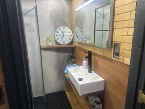A bathroom with a white sink, black faucet, and two clocks on the wall. There is a mirror above the sink and a small shelf with plants. - Stunning 1-bed Studio in London (London)