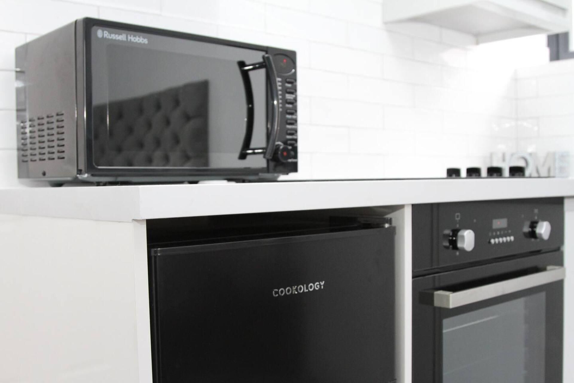 Une cuisine avec un micro-ondes noir posé sur un plan de travail blanc, un réfrigérateur noir sur lequel on peut lire le mot « COOKOLOGY », et un four encastré.