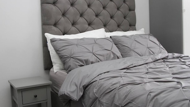 Ein Bett mit einer gesteppten Kopfteil, grauer Bettwäsche und einem kleinen Nachttisch mit Schublade.