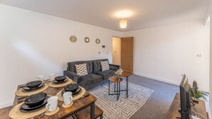 Een moderne woonkamer met een bank, een eettafel gedekt met borden en bekers, een klein bijzettafeltje met een plant en een plafondlamp.