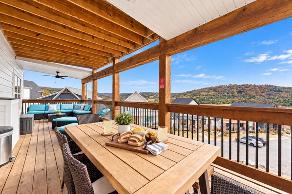 A wooden deck with outdoor furniture, a table with food and drinks, and a view of a hilly landscape.