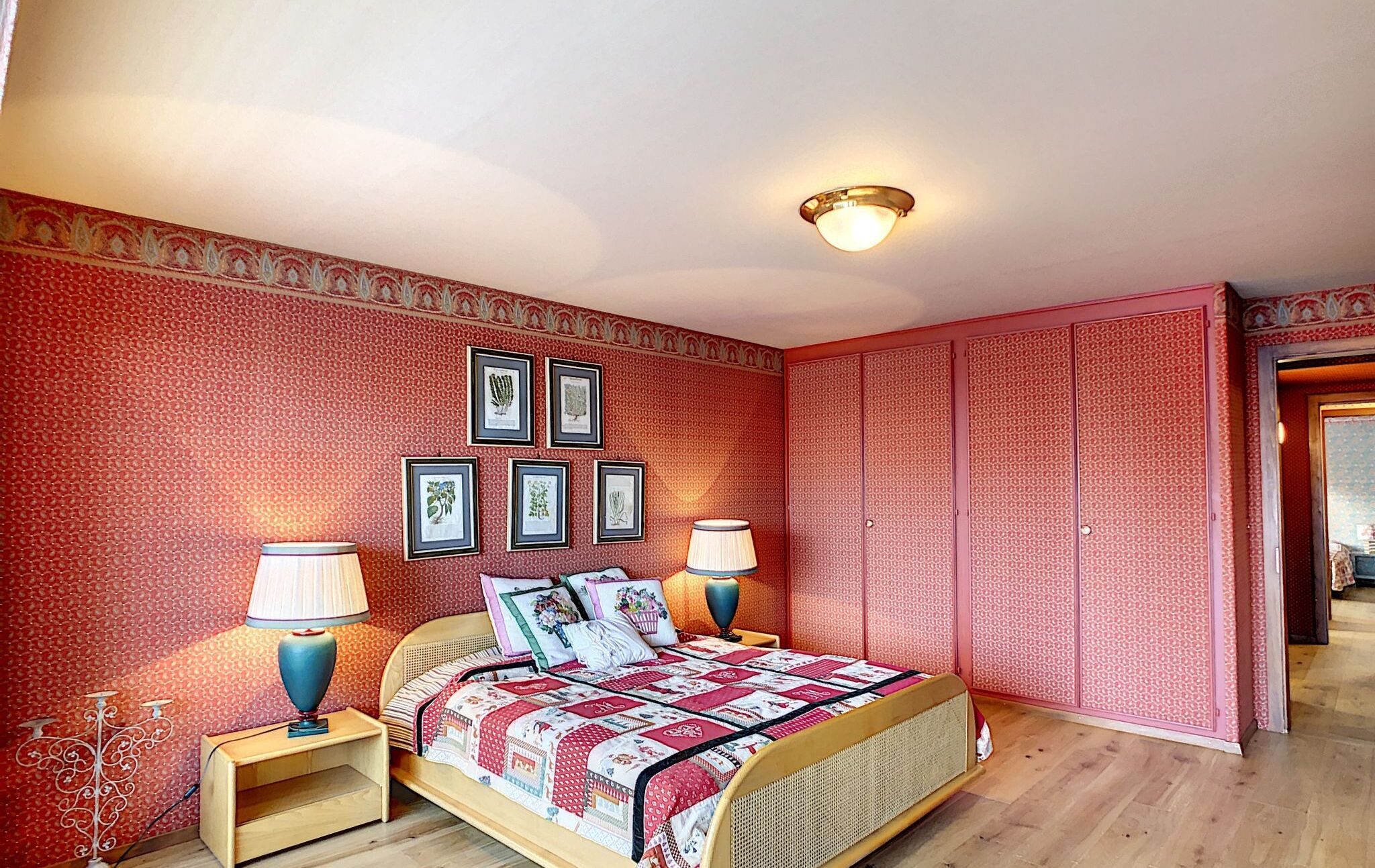 Ein Schlafzimmer mit strukturiertem Wandpapier, einem Bett mit Patchworkdecke, zwei Nachttischlampen und gerahmten Pflanzenbildern an der Wand.