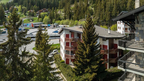 A mountain resort with a parking lot, buildings with balconies, and a forested backdrop.