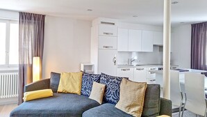 A modern living room with a grey sofa, patterned cushions, and a small white side table. A kitchen with white cabinets and a dining area with a white table and chairs is visible in the background.