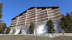 Un hotel de varios pisos con balcones, un muro de piedra de contención y árboles alrededor de la propiedad.