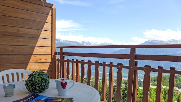 Una mesa redonda con una revista, un vaso con forma de corazón y una planta pequeña en un balcón con barandilla de madera, con vistas a un paisaje montañoso.