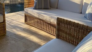 A sunlit outdoor seating area with a wicker sofa, cushions, and a view of a mountain range.