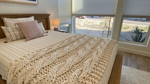 A bedroom with a large bed, a headboard, a nightstand, a lamp, and a window with a view of a landscape.