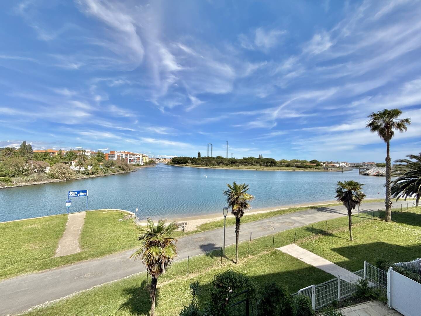 A riverside view with a clear sky, palm trees, and a pathway leading to a building.