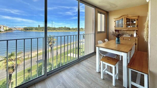 Un balcon donnant sur une rivière et des immeubles, une table en bois avec des chaises, et un meuble avec des portes en verre.