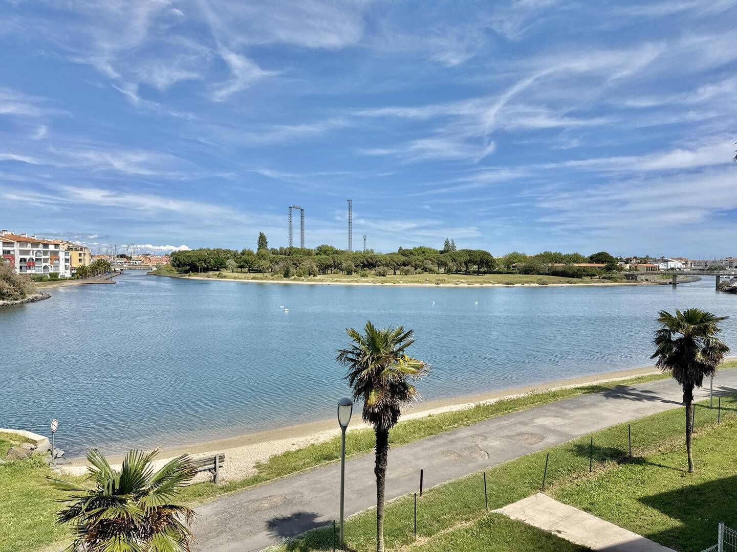 A river with a pedestrian path, palm trees, and buildings in the background.