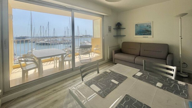 A room with a large sliding glass door leading to a balcony with a marina view. Inside, there is a dining table with placemats and a sofa. - CAP D'AGDE - L'Ile des pêcheurs / Réf 0216 (CAP D'AGDE)