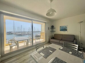A room with a large sliding glass door leading to a balcony with a marina view. Inside, there is a dining table with placemats and a sofa. - CAP D'AGDE - L'Ile des pêcheurs / Réf 0216 (CAP D'AGDE)