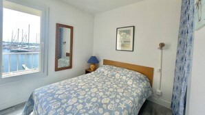 A bedroom with a bed, a window with a view of boats, a framed picture on the wall, and a lamp on a bedside table. - CAP D'AGDE - L'Ile des pêcheurs / Réf 0216 (CAP D'AGDE)