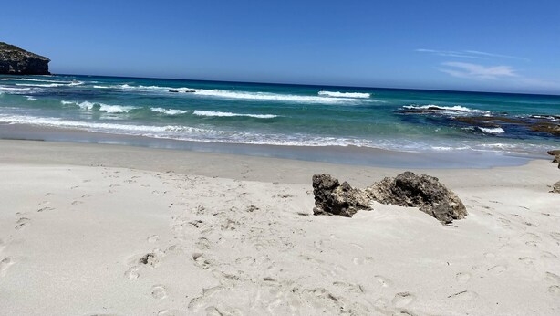 Una playa con agua azul cristalina, orilla arenosa y afloramientos rocosos.