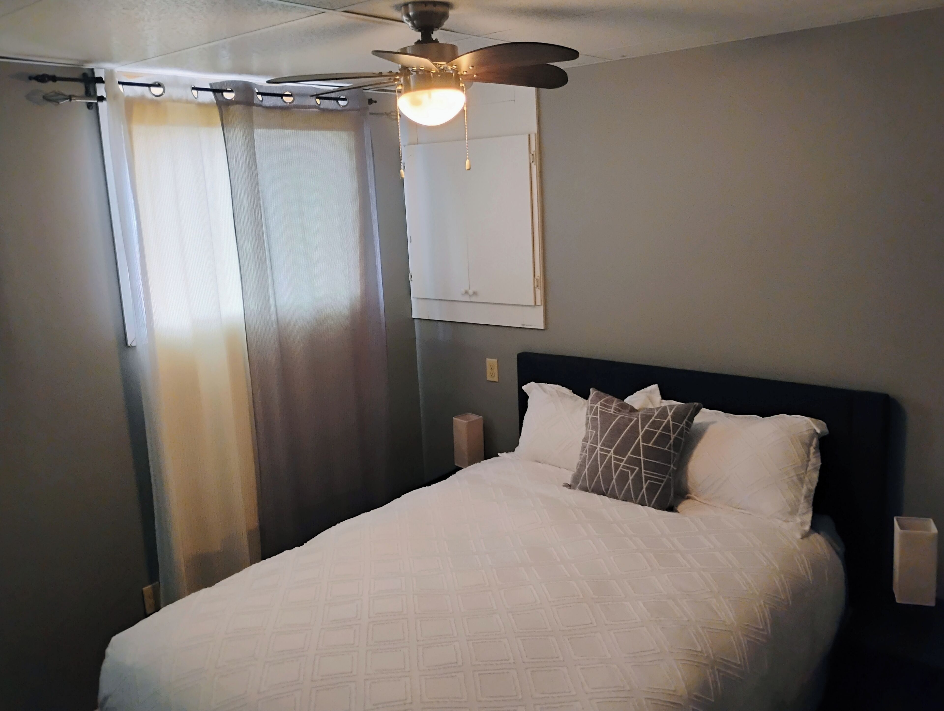 A hotel room with a bed, a ceiling fan, a window with curtains, and a wall-mounted light switch.