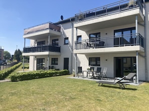 Um edifício residencial de dois andares com varandas, um espaço de pátio com mobiliário exterior e um gramado bem cuidado.