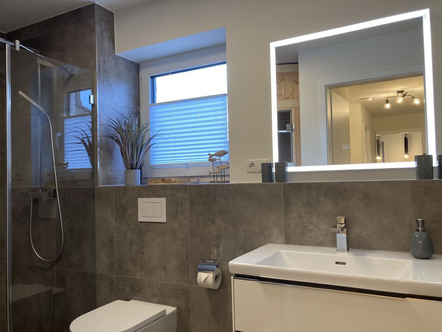A modern bathroom with a glass shower enclosure, a toilet, a sink with a mirror, and a window with blinds.