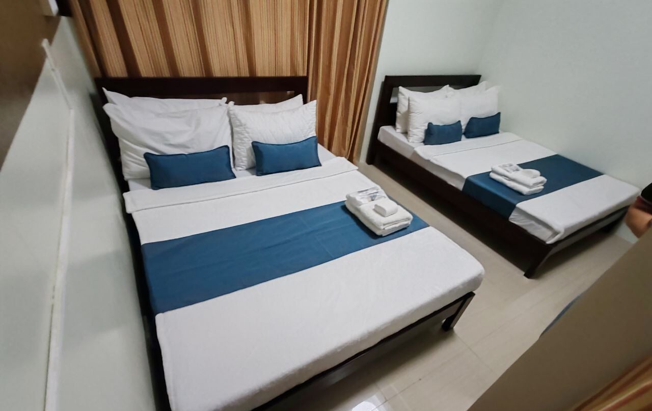 Dos camas individuales con ropa de cama blanca y azul, un cabecero de madera y una cortina.
