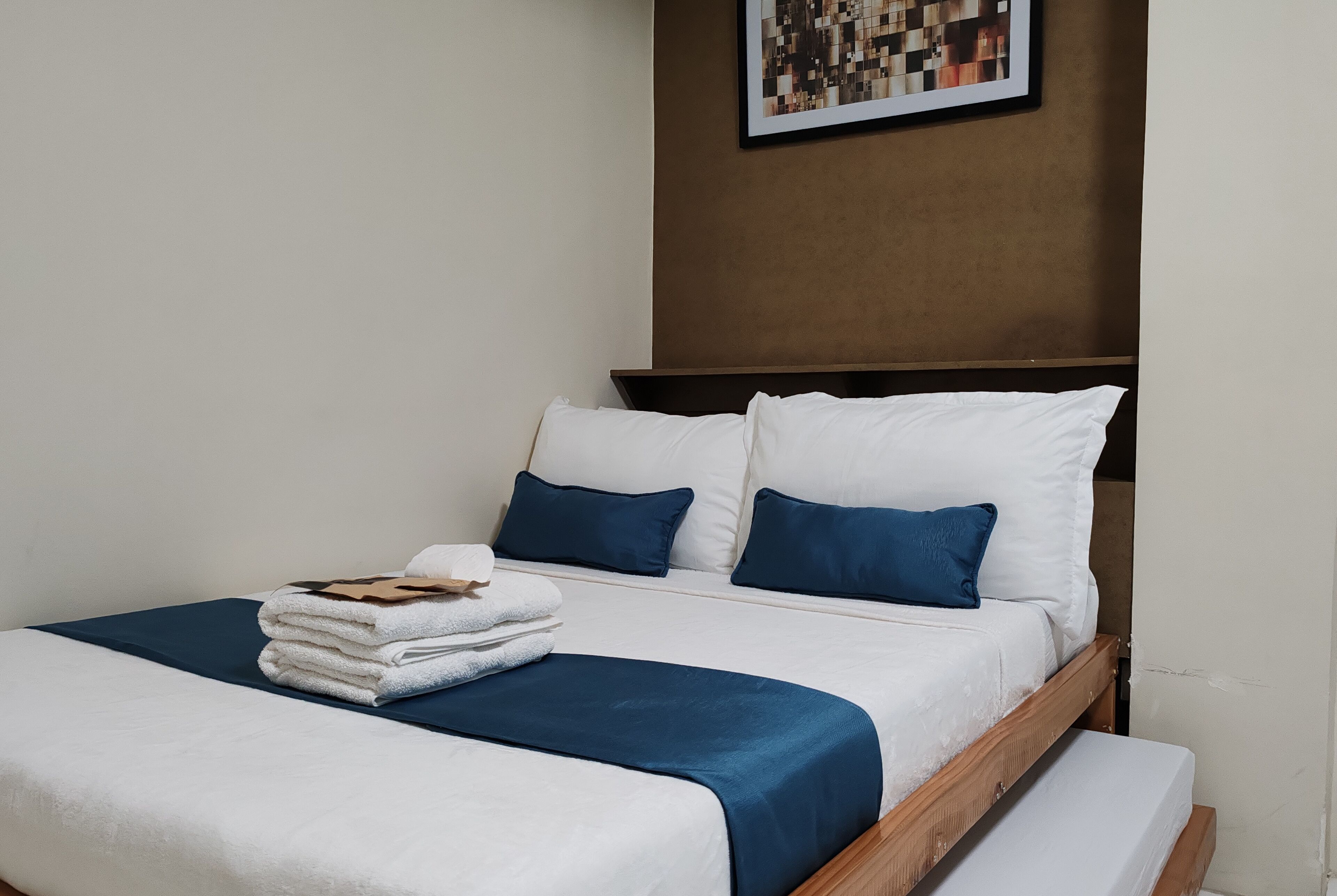 A neatly made bed with white and blue bedding, a stack of folded towels, and a framed picture on the wall.